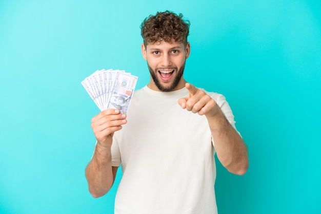 Jeune homme caucasien prenant beaucoup d'argent isolé sur fond bleu surpris et pointant vers l'avant