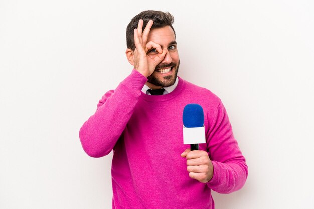 Jeune homme caucasien présentateur de télévision isolé sur fond blanc excité en gardant le geste ok sur les yeux