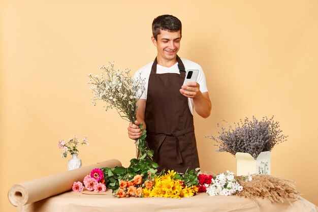 Jeune homme caucasien propriétaire d'un centre de fleurs en tapant sur le téléphone responsable des employés en tablier faisant l'inventaire homme gai utilisant un smartphone dans un magasin de fleurs isolé sur fond beige