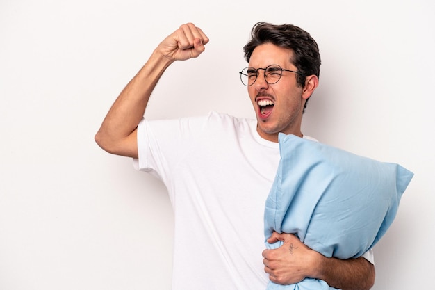 Jeune homme caucasien en pyjama tenant un oreiller isolé sur fond blanc levant le poing après un concept de vainqueur de la victoire