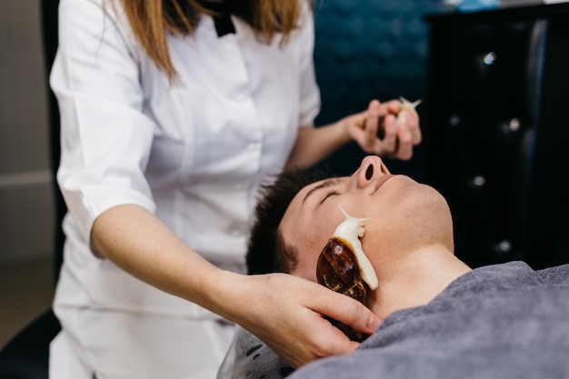 Jeune homme caucasien recevant un massage aux escargots Thérapie et soins de la peau du visage avec des escargots dans un salon de spa Procédure cosmétologique moderne de rajeunissement de la peau