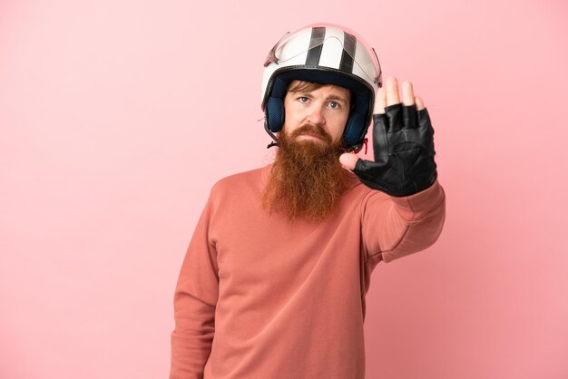 Jeune homme caucasien rougeâtre avec un casque de moto isolé sur fond rose faisant un geste d'arrêt