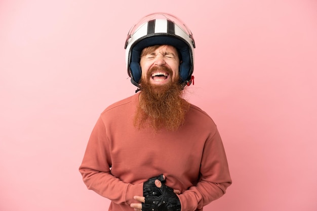 Jeune homme caucasien rougeâtre avec un casque de moto isolé sur fond rose souriant beaucoup