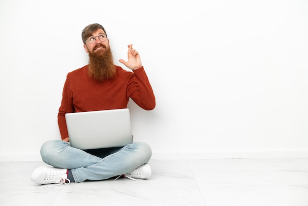 Jeune homme caucasien rougeâtre avec ordinateur portable isolé sur fond blanc avec les doigts croisés et souhaitant le meilleur