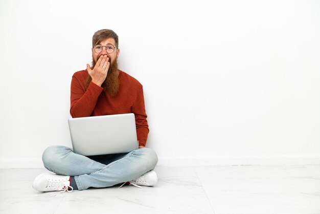 Jeune homme caucasien rougeâtre avec ordinateur portable isolé sur fond blanc heureux et souriant couvrant la bouche avec la main