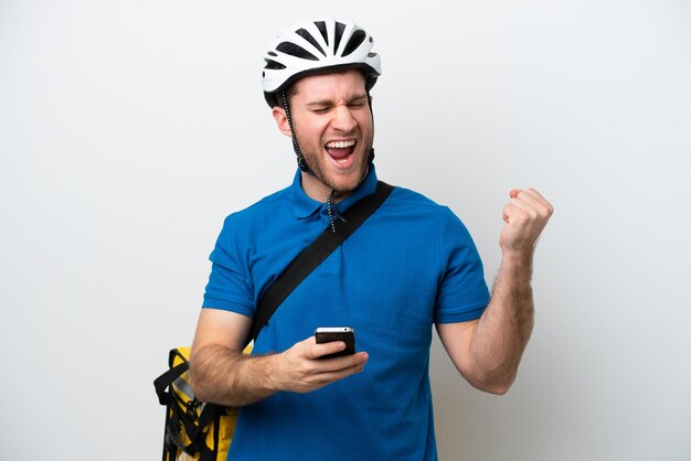 Jeune homme caucasien avec sac à dos thermique isolé sur fond blanc avec téléphone en position de victoire