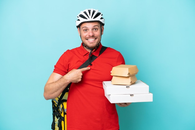 Jeune homme caucasien avec sac à dos thermique et tenant de la restauration rapide isolé sur fond bleu avec une expression faciale surprise