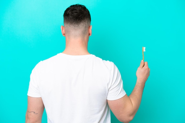 Jeune homme caucasien se brosser les dents isolé sur fond bleu en position arrière