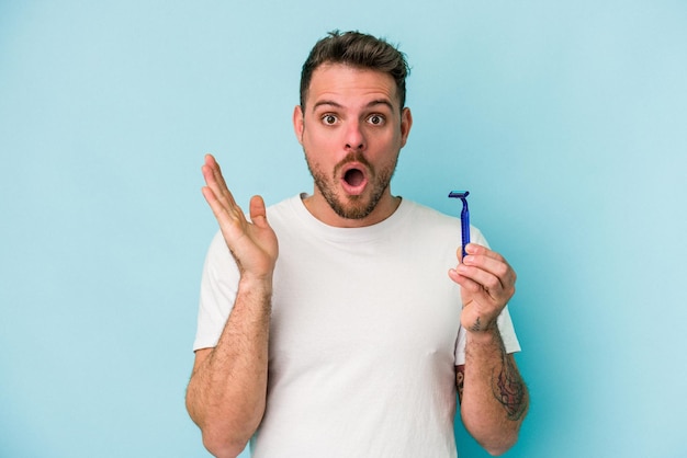 Jeune homme caucasien se rasant la barbe isolé sur fond bleu surpris et choqué