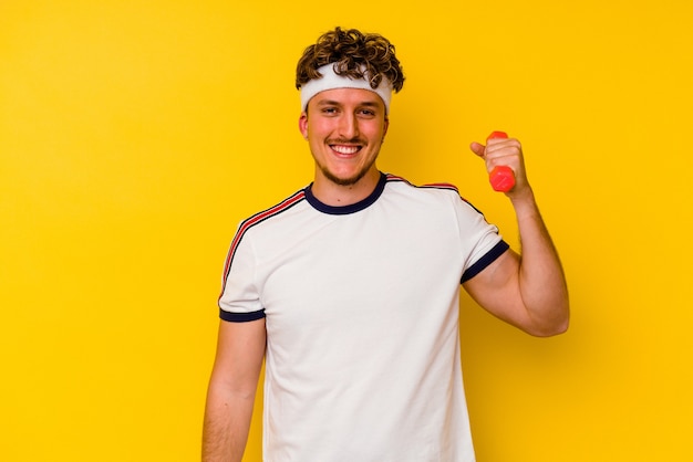 Jeune homme caucasien de sport tenant un haltère isolé sur fond jaune heureux, souriant et joyeux.
