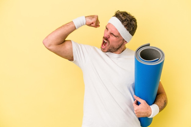 Jeune homme caucasien sportif allant à des cours de yoga tout en tenant un tapis isolé sur fond jaune levant le poing après une victoire, concept gagnant.