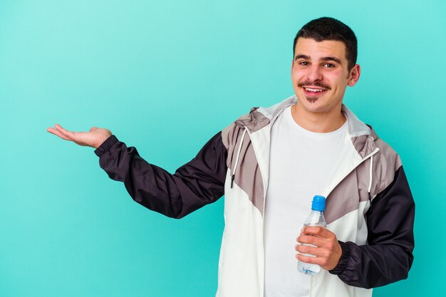 Jeune homme caucasien sportif eau potable isolée sur bleu montrant un espace de copie sur une paume et tenant une autre main sur la taille.