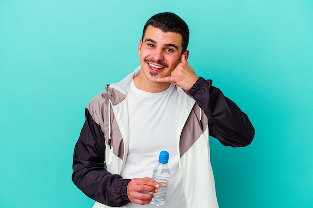 Jeune homme caucasien sportif eau potable isolée sur bleu montrant un geste d'appel de téléphone mobile avec les doigts.