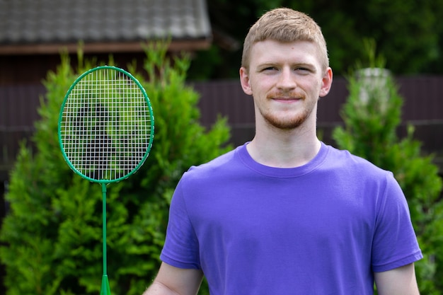 Jeune homme caucasien sportif fort posant avec une raquette de badminton sur la nature verte. concept de jeu amateur de badminton, activités de plein air d'été.