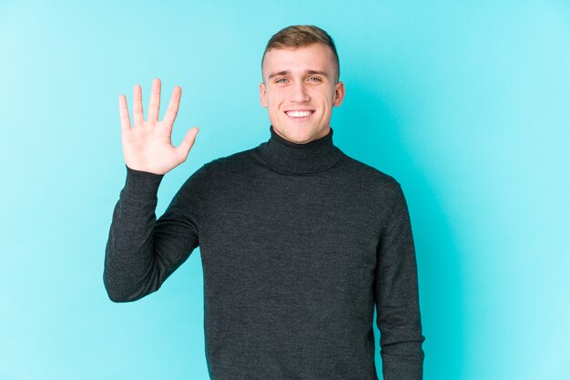 Jeune homme caucasien sur une surface bleue souriant joyeux montrant le numéro cinq avec les doigts.