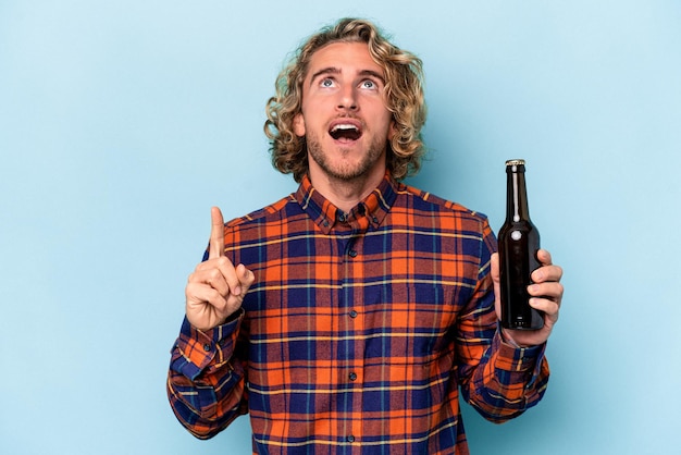 Jeune homme caucasien tenant une bière isolée sur fond blanc pointant vers le haut avec la bouche ouverte.
