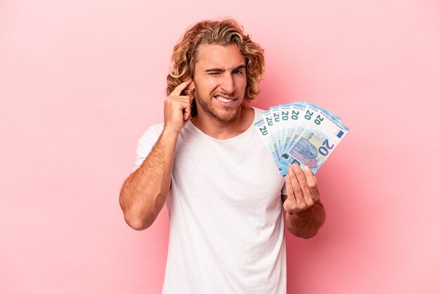 Jeune homme caucasien tenant des billets de banque isolés sur fond rose couvrant les oreilles avec les mains.