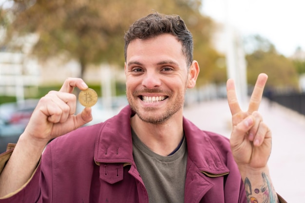 Jeune homme caucasien tenant un Bitcoin à l'extérieur souriant et montrant le signe de la victoire