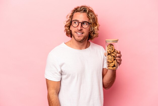 Jeune homme caucasien tenant un bocal à biscuits isolé sur fond rose regarde de côté souriant, joyeux et agréable.