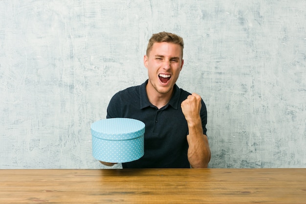 Jeune homme caucasien tenant une boîte-cadeau sur une table applaudissant sans soucis et excité. Concept de victoire.