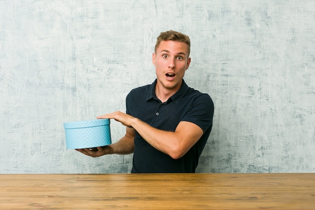 Jeune homme caucasien tenant une boîte-cadeau sur une table choqué et étonné tenant un espace de copie entre les mains.