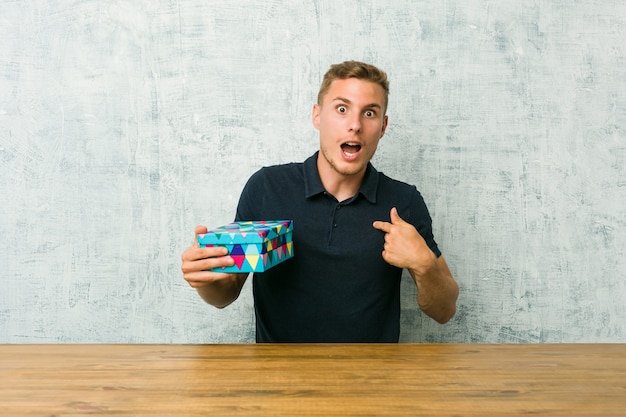 Jeune homme caucasien tenant une boîte-cadeau sur une table surpris en pointant avec le doigt, souriant largement.