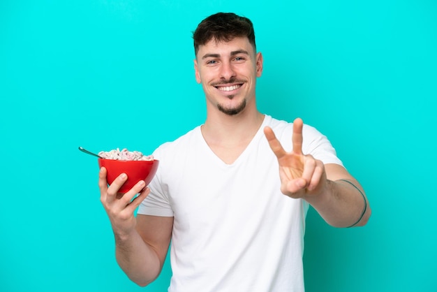 Jeune homme caucasien tenant un bol de céréales isolé sur fond bleu souriant et montrant le signe de la victoire