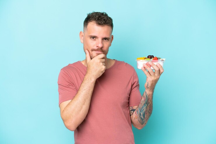 Photo jeune homme caucasien tenant un bol de fruits isolé sur fond bleu pensant