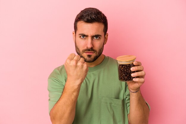 Jeune homme caucasien tenant une bouteille de café isolée sur fond rose montrant le poing à la caméra, expression faciale agressive.