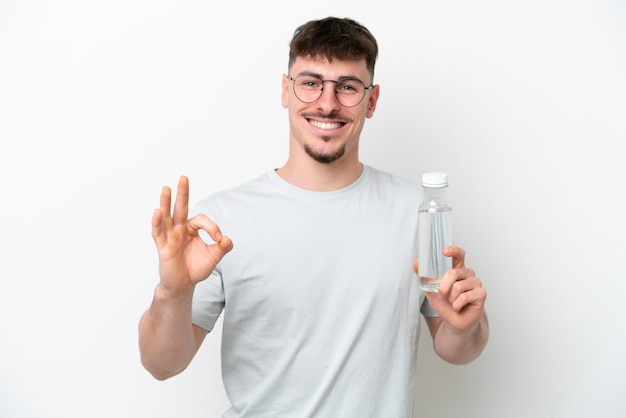 Jeune homme caucasien tenant une bouteille d'eau isolé sur fond blanc montrant signe ok avec les doigts