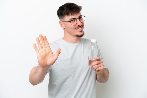 Jeune homme caucasien tenant une bouteille d'eau isolé sur fond blanc saluant avec la main avec une expression heureuse