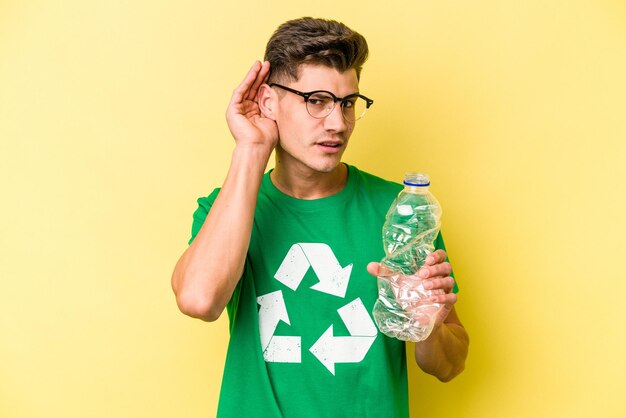 Jeune homme caucasien tenant une bouteille de plastique à recycler isolé sur fond jaune essayant d'écouter un potin.