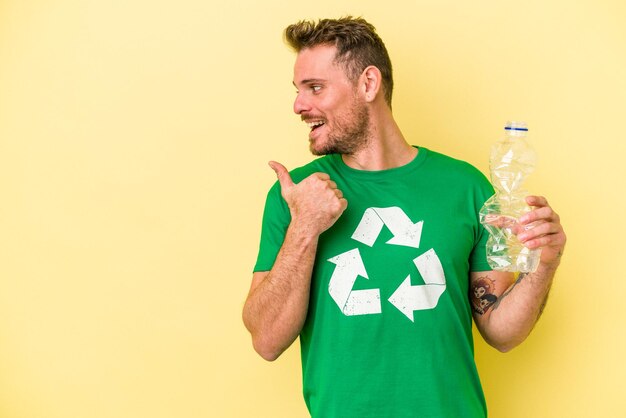 Jeune homme caucasien tenant une bouteille de plastique à recycler isolé sur fond jaune pointe avec le pouce loin, riant et insouciant.