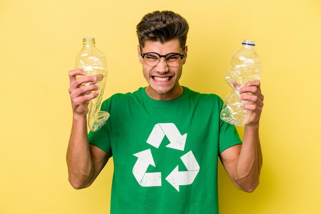 Jeune homme caucasien tenant une bouteille de plastique à recycler isolé sur fond jaune