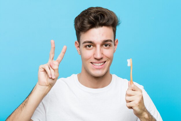 Jeune homme caucasien tenant une brosse à dents Jeune femme indienne portant un pyjama et un masque de sommeil isolé tenant un oreiller montrant le signe de la victoire et souriant largement. <Mixto>