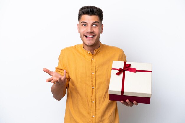 Jeune homme caucasien tenant un cadeau isolé sur fond blanc avec une expression faciale choquée