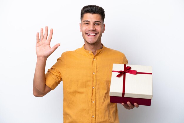 Jeune homme caucasien tenant un cadeau isolé sur fond blanc saluant avec la main avec une expression heureuse