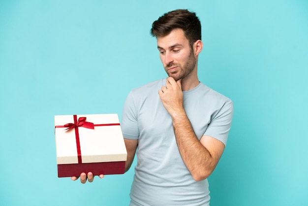 Jeune homme caucasien tenant un cadeau isolé sur fond bleu avec une expression triste