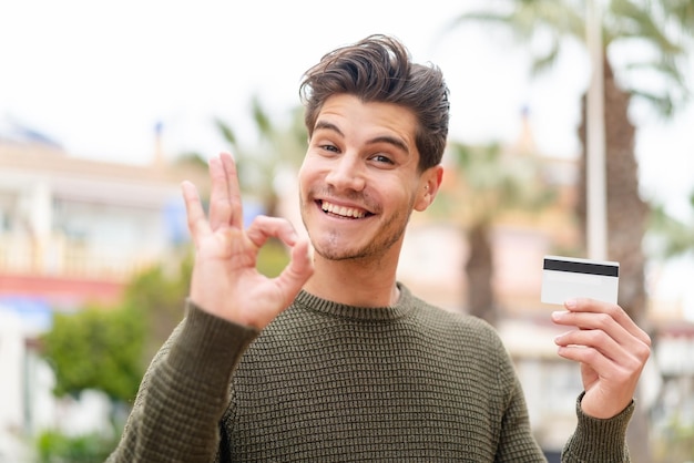 Jeune homme caucasien tenant une carte de crédit à l'extérieur montrant un signe ok avec les doigts