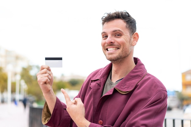 Jeune homme caucasien tenant une carte de crédit à l'extérieur et la pointant