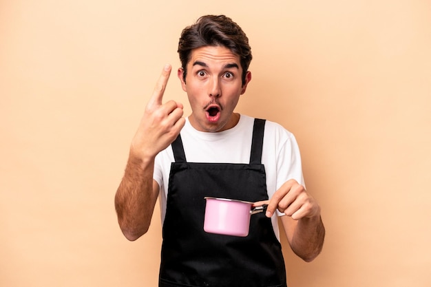 Jeune homme caucasien tenant une casserole isolée sur fond beige ayant un concept d'inspiration idée