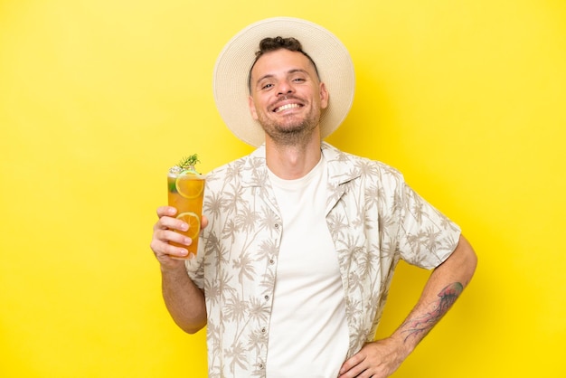 Jeune homme caucasien tenant un cocktail isolé sur fond jaune posant avec les bras à la hanche et souriant