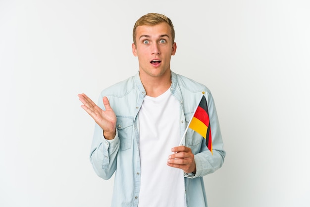 Jeune homme caucasien tenant un drapeau allemand isolé sur un mur blanc surpris et choqué.