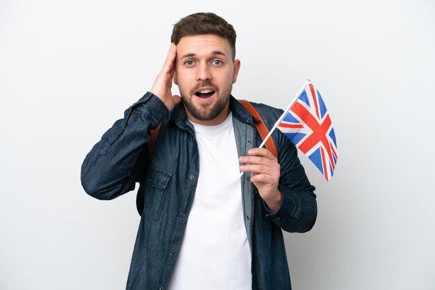 Jeune homme caucasien tenant un drapeau du Royaume-Uni isolé sur fond blanc avec une expression de surprise