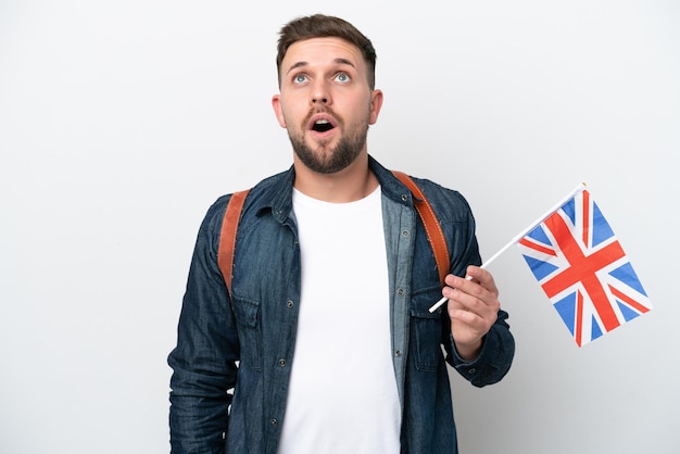 Jeune homme caucasien tenant un drapeau du Royaume-Uni isolé sur fond blanc en levant et avec une expression surprise