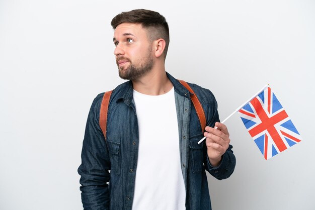 Jeune homme caucasien tenant un drapeau du Royaume-Uni isolé sur fond blanc levant les yeux en souriant