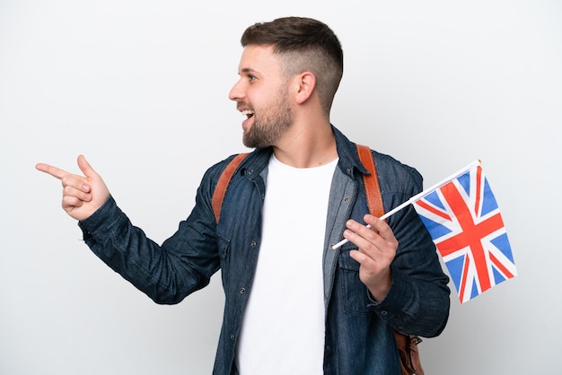 Jeune homme caucasien tenant un drapeau du Royaume-Uni isolé sur fond blanc pointant le doigt sur le côté et présentant un produit