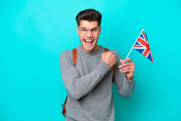 Jeune homme caucasien tenant un drapeau du Royaume-Uni isolé sur fond bleu célébrant une victoire