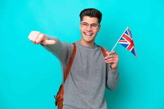 Jeune homme caucasien tenant un drapeau du Royaume-Uni isolé sur fond bleu donnant un geste du pouce levé