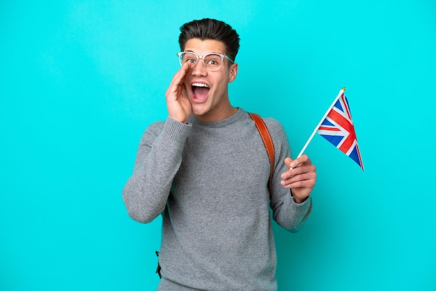 Jeune homme caucasien tenant un drapeau du Royaume-Uni isolé sur fond bleu avec une expression faciale surprise et choquée